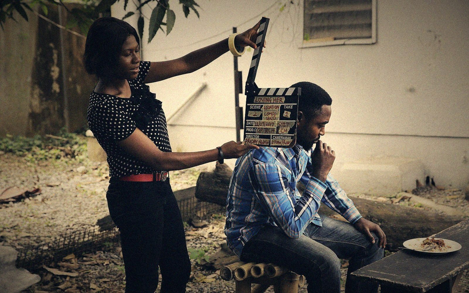 A woman holds a clapperboard beside a seated actor during an outdoor film shoot