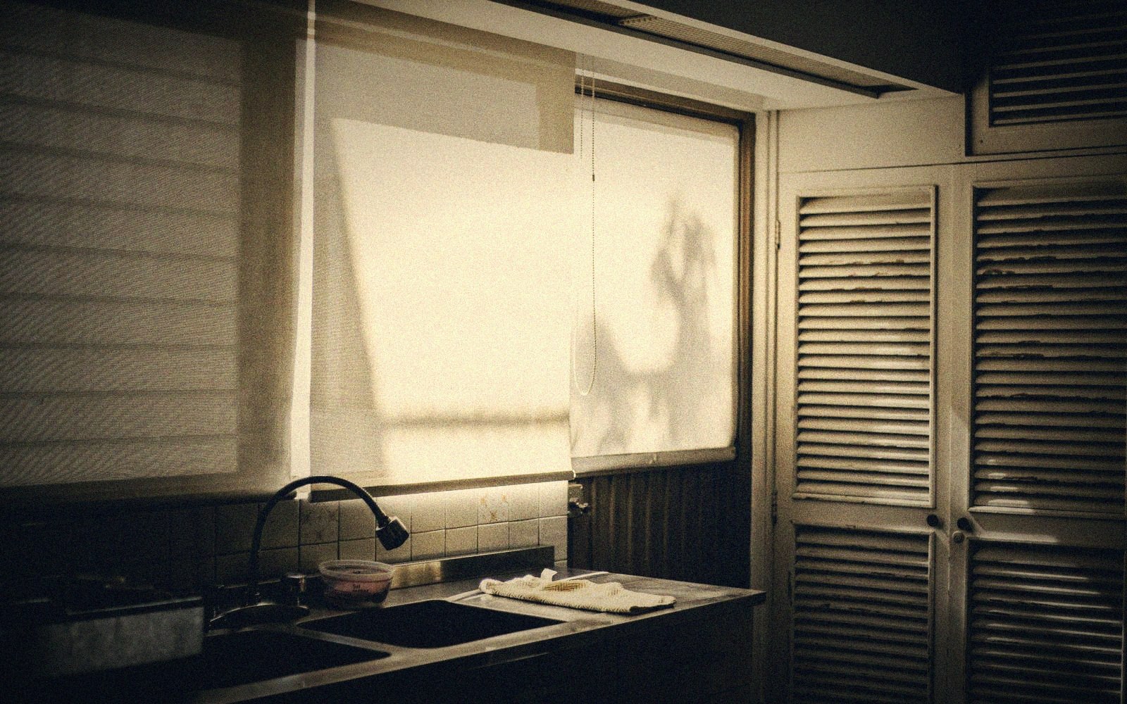 Sunlit roller blinds cast shadows above a kitchen sink beside louvered cabinet doors