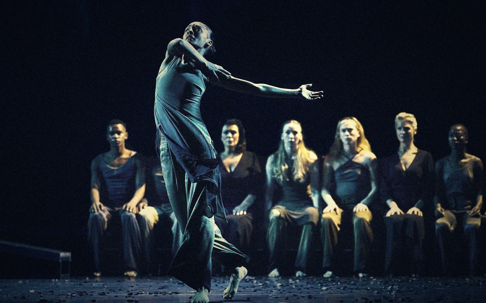 A dancer reaches forward mid-movement while seated performers watch from chairs onstage
